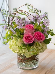 a glass vase filled with pink roses and greenery on top of a wooden table