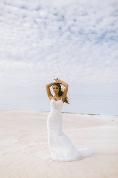 a woman in a white dress standing on the beach with her hands behind her head