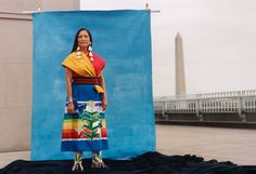 a woman standing in front of a blue backdrop wearing a colorful skirt and yellow shawl