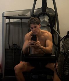 a shirtless man sitting in a chair looking at his cell phone while holding a fan