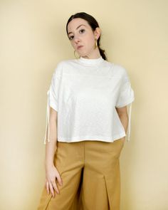 a woman standing in front of a wall wearing brown pants and a white top with fringes