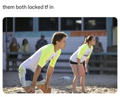 two people in yellow shirts are playing frisbee on the beach while others watch