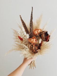 a hand is holding a dried flower arrangement with feathers and flowers in it's centerpiece