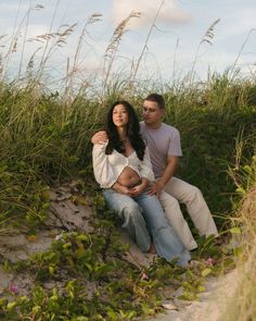 a man and woman sitting on top of a sandy beach next to tall green grass