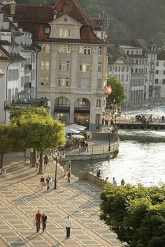 several people walking on the sidewalk in front of some buildings and water with mountains in the background