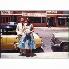 a man and woman standing in front of an old car on the side of the road