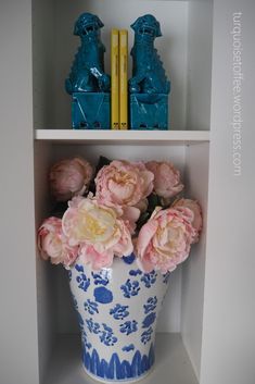 a blue and white vase filled with pink flowers on top of a book shelf next to two yellow books