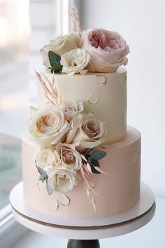 a three tiered cake with white and pink flowers on the top, next to a window