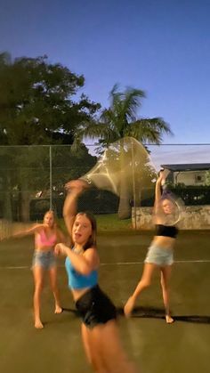 three girls are playing tennis on the court at night, and one girl is reaching up to hit the ball with her racquet