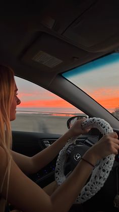 a woman driving a car with the sun setting in the distance behind her and she's wearing a white knitted steering wheel cover