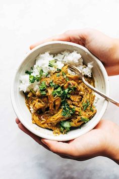 two hands holding a bowl of food with rice and meat in it on a white surface