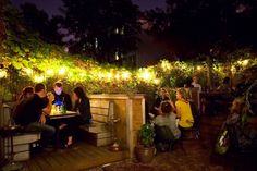 a group of people sitting around a table outside at night with lights on the trees