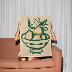 a woman sitting on a couch holding up a painting with lemons in a bowl