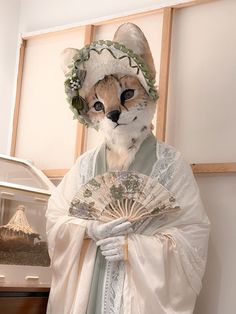 a cat wearing a costume and holding a fan in front of a wall with pictures on it