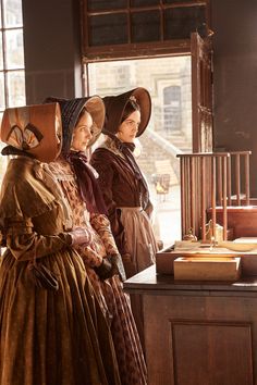 two women dressed in period clothing standing next to each other and looking out the window