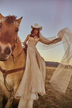 a woman in a white dress is standing next to a brown horse and wearing a cowboy hat