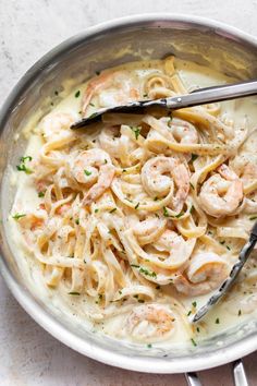 pasta with shrimp and cream sauce in a silver bowl on a white tablecloth next to two serving utensils