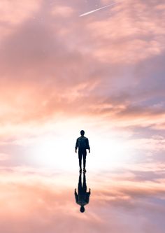 a man standing in the middle of a body of water under a pink sky with an airplane flying overhead