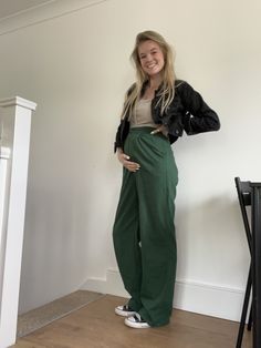 a woman standing in front of a white wall wearing green pants and a black jacket