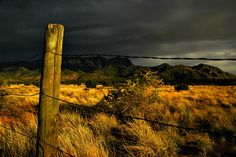 a fence in the middle of a field with mountains in the background and dark clouds overhead