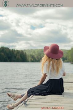 a woman sitting on top of a wooden pier near the water wearing a red hat