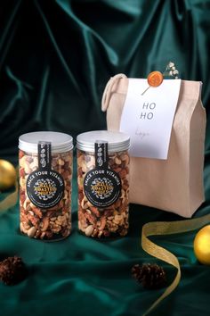 two jars filled with nuts sitting next to a brown paper bag and gold ornaments on a green cloth