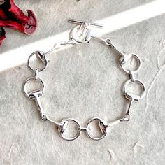 Inspired by equestrian elegance, this Snaffle Bit Chunky Sterling Silver Charm Bracelet features distinctive horse-bit charms and a functional t-bar fastening.  Hand-assembled with precision, each silver link creates a timeless, dramatic statement piece that's both sophisticated and versatile. Perfect for daily wear or as a treasured heirloom, this bracelet makes a thoughtful gift for horse lovers and those who appreciate bold, elegant jewelry. Made From: Sterling Silver Measurements: Bracelet l Chunky Silver Bracelet, Snaffle Bit, Horse Jewelry, Sterling Silver Charm Bracelet, Horse Lovers, Gifts For Horse Lovers, Silver Charm Bracelet, Statement Bracelet, Bracelet Silver