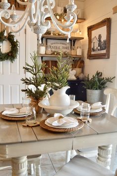 a dining room table set for christmas with white dishes and greenery in the center