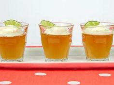 three shot glasses filled with orange juice and limes on a red tablecloth covered serving tray