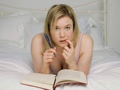 a woman laying in bed with an open book and pen looking at the camera while reading