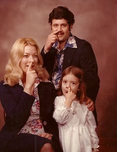 a man and woman pose for a photo with their daughter, who is holding her finger to her mouth