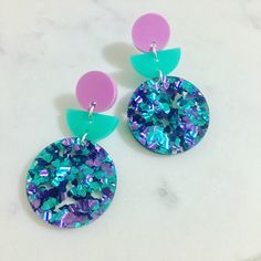 two pairs of purple, green and blue earrings on a marble surface with pink circles