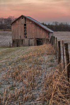 an old barn sits in the middle of a field at sunset with frost on the grass