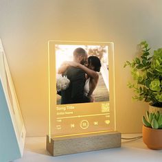 an illuminated photo frame sitting on top of a desk next to a potted plant