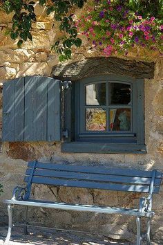 a blue bench sitting in front of a window with shutters open and flowers hanging over it