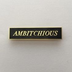 a black and gold pin with the words ambitioushouse on it's back side