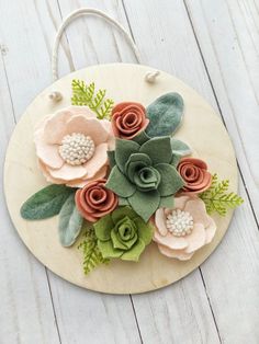 a wooden plaque with felt flowers and greenery on the front, sitting on a white wood background