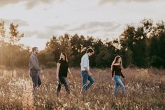 four people are walking through tall grass in the sunlit field with trees behind them