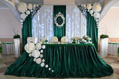 the table is set with white flowers and greenery on it, along with green drapes