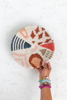 a hand holding a colorful woven basket on top of a white wall next to a stack of bracelets