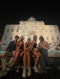 four people are sitting on a bench in front of a building at night, posing for the camera