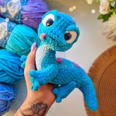 a hand holding a blue crocheted toy in front of balls of yarn and flowers