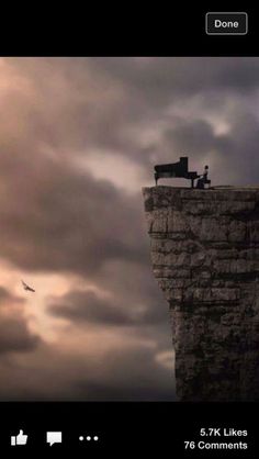 a man sitting on top of a cliff next to a black and white bench under a cloudy sky