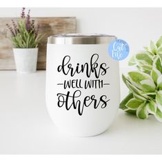 a white cup with the words drinks well with others on it next to some plants