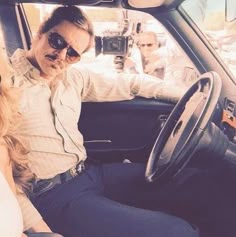 a man and woman sitting in the driver's seat of a car with cameras around them