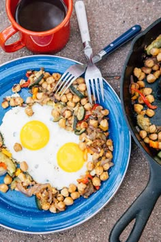 fried eggs and vegetables on a blue plate next to a cup of coffee, fork and spoon