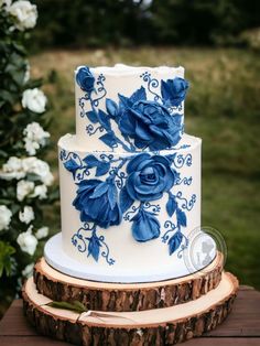 a white and blue wedding cake with roses on it sitting on a tree stump in front of flowers