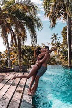 a man and woman standing on the edge of a swimming pool with palm trees in the background