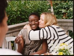 two women embracing each other while sitting at a table outside with flowers and greenery in the background