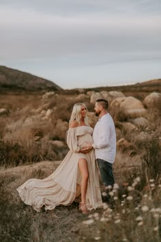 a man and woman standing next to each other in the grass with rocks behind them